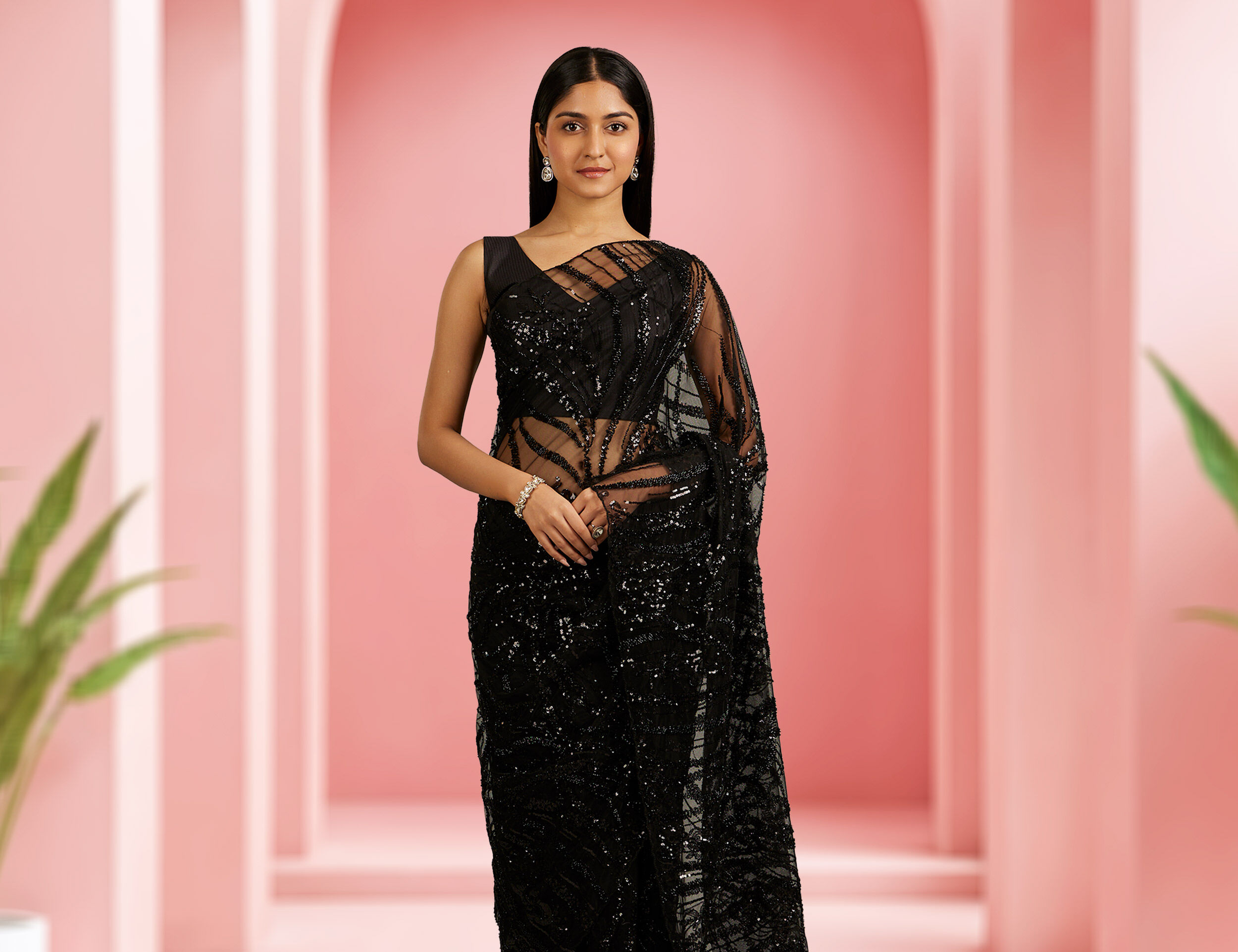 Woman in a black sequined saree with sheer panel blouse, styled with minimal jewelry, standing against a soft pink arched backdrop